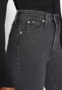 Höga svarta jeans med klassisk passform, med silverdetaljer, subtila sömmar och en mjuk denimtextur. CK-logotyp på midjebandet.