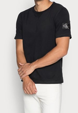 Schwarzes T-Shirt mit kurzen Ärmeln aus Baumwolle, mit Rundhals und einem Markenaufnäher mit Initialen am linken Ärmel. Normale Passform.