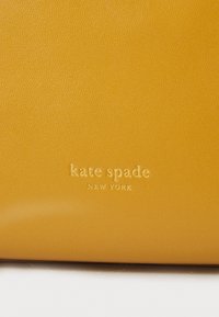 Keltainen nahkabag, jossa on kohokuvioitu "kate spade NEW YORK" -logo. Tasainen pinta, jossa on hieman kiiltoa ja jämäkkä muotoilu.