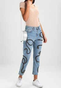 Lysvaskede denimjeans med et design af en snoet slange i sort med hvide detaljer. Kombinationen består af en tætsiddende beige top og en rund sølvfarvet taske.