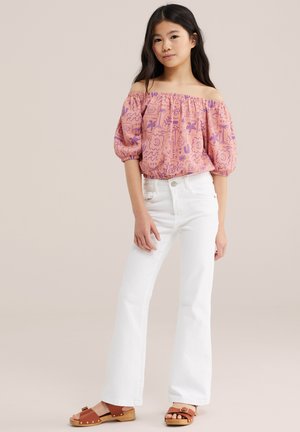 Lány rózsaszín váll nélküli blúzban, lila mintákkal, fehér bő szárú nadrágban és barna szandálban, bézs háttér előtt áll.