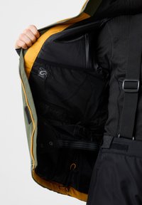 Grön ytterjacka med en kontrasterande gul inredning. Har en svart meshficka, justerbara remmar och dragkedjedetaljer.