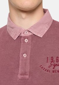 Koszulka polo z krótkim rękawem w średnim odcieniu śliwkowym, z teksturowanej tkaniny, jaśniejszym kołnierzykiem, trzema guzikami i haftowanymi detalami.