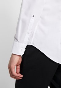 Witte blouse met lange mouwen en een puntige kraag. Voorzien van zwarte rozenborduursels op de manchetten en witte knopen. Glad textuur van de stof.