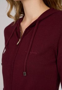 Haut à capuche zippé bordeaux en tissu doux. Équipé d'un cordon de serrage avec des accents métalliques et d'un logo brodé subtil sur la poitrine.