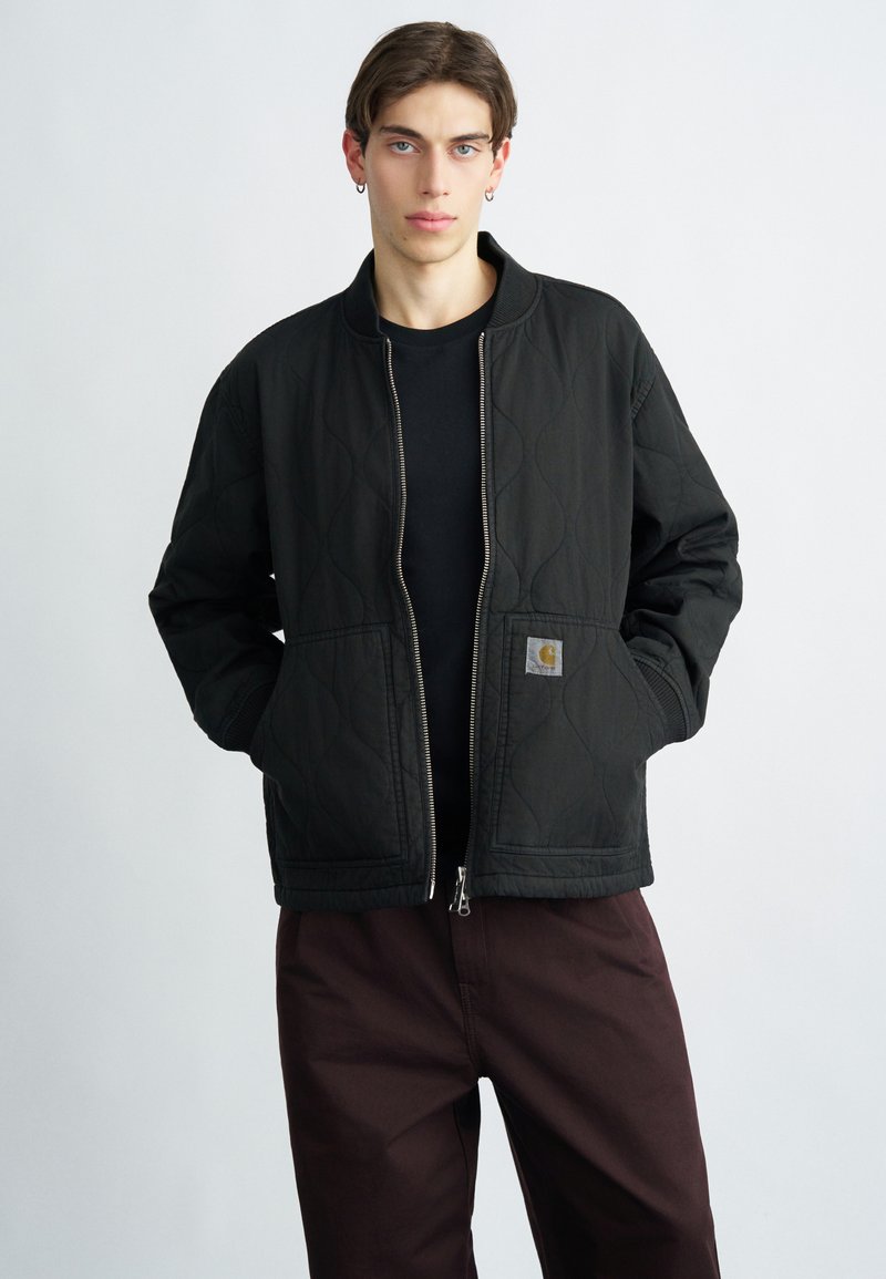 Black quilted jacket with a high collar, silver zipper, and a patch logo on the left pocket. Paired with dark brown pants.