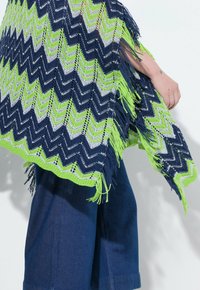 Virkat poncho i marinblått, grönt och vitt zigzag-mönster, med fransar i kanten. Kombinerat med denimbyxor. Strukturerad, lättviktsmaterial.