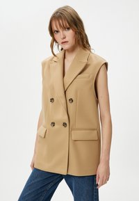 Gilet beige senza maniche doppiopetto in morbido tessuto, con due tasche frontali e bottoni tondi, abbinato a pantaloni in denim blu.