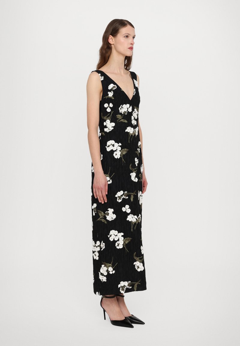Robe noire à fleurs avec des fleurs blanches brodées et des feuilles vertes, présentant un décolleté en V profond et une silhouette fluide. Associée à des talons noirs.