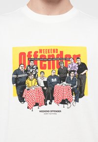 White t-shirt featuring a bright yellow and red graphic with a group of men seated at checkered tables, labeled "WEEKEND OFFENDER."