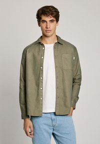 Camicia verde oliva con bottoni, dotata di una tasca sul petto, abbinata a una maglietta bianca e jeans azzurri chiari. Tessuto morbido, vestibilità rilassata.