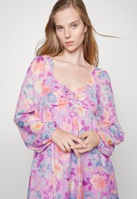 Vestido floral em tecido suave e transparente com uma paleta de cores roxa e pastel. Apresenta um decote quadrado, detalhe franzido na frente e mangas longas bufantes.