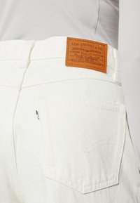 Biele džíny z denimu s hnedým koženým štítkom na zadnej strane, na ktorom je logo Levi Strauss a malá nálepka s podrobnosťami o značke.