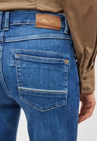 Τζιν παντελόνια σε μεσαίο μπλε χρώμα με καφέ δερμάτινη ετικέτα, που διαθέτουν μία πίσω τσέπη με άσπρες ραφές ως διακοσμητικό στοιχείο.