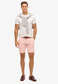 Pantalones cortos rosa claro, camiseta blanca, suéter gris colgado sobre los hombros, cinturón marrón y zapatos de barco color burdeos. Tejido suave y corte ajustado.