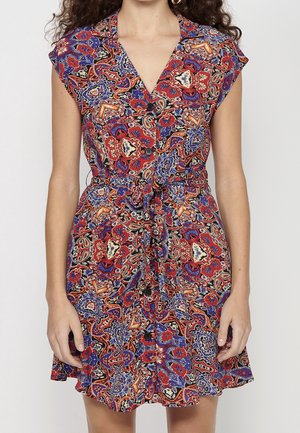 Frau trägt ein ärmelloses, geknöpftes Kleid mit einem bunten roten, blauen und schwarzen Blumen- und Paisley-Muster und einem gebundenen Taillengürtel.