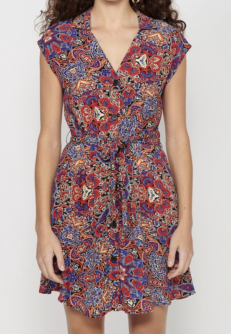 Femme portant une robe sans manches boutonnée avec un motif floral et cachemire coloré rouge, bleu et noir, et une ceinture nouée à la taille.