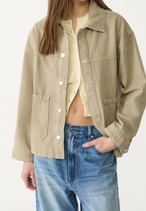 Mujer con chaqueta beige abotonada sobre top amarillo corto y vaqueros azules de talle alto, mano en el bolsillo, recortada en cuello y caderas.