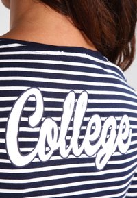 Marinblå och vit randig t-shirt med ordet "College" i stora, vita, stiliserade bokstäver på baksidan. Tillverkad i bomull, avslappnad design.