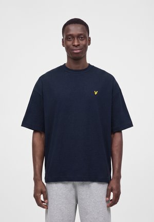 Ung mand iført en løs mørkeblå t-shirt med et lille gult fuglelogo på brystet og lysegrå bukser, stående mod en ensfarvet baggrund.