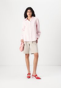 Camicia a righe rosa con bottoni, pantaloni corti beige su misura, borsa a tracolla rosa, scarpe con tacco rosse con accenti metallici. Texture lisce, vestibilità casual.