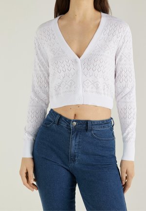 Femme portant un cardigan blanc court boutonné avec un tricot à motif losange et un jean bleu taille haute sur un fond neutre.