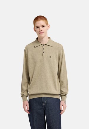 Giovane uomo con capelli rossi corti che indossa un maglione polo beige a maniche lunghe e jeans blu scuro in piedi davanti a uno sfondo semplice.