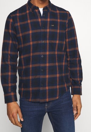 Hombre con una camisa de botones de manga larga a cuadros azul marino y naranja óxido con vaqueros azules.