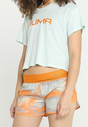 Kobieta nosząca jasnoniebieski krótki t-shirt Puma oraz sportowe spodenki z pomarańczowymi akcentami i wzorzystym designem, stoi na tle jednolitego tła.