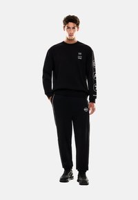 Černá mikina s logem a textem na hrudi a rukávu, doplněná černými tepláky s logem. Nosit s černými teniskami.