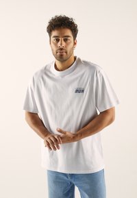 White cotton t-shirt featuring short sleeves and a round neck. Includes a small graphic in blue text on the left chest area. Casual fit.