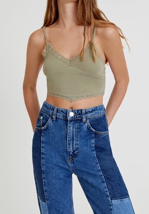 Femme portant un crop top vert orné de dentelle et un jean patchwork en denim bleu taille haute, debout les mains dans les poches.