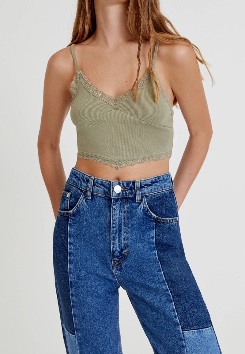 Femme portant un crop top vert orné de dentelle et un jean patchwork en denim bleu taille haute, debout les mains dans les poches.