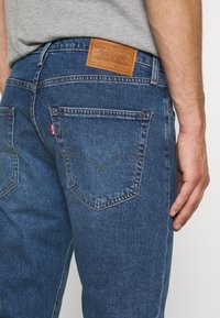 Blå denimjeans med en brun läderlapp på baksidan av midjan, synlig söm och två bakfickor med en klassisk design.