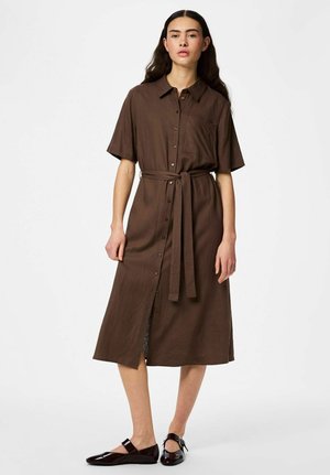 Kvinde iført en brun midi-kjole med korte ærmer, knapper foran og bælte i taljen samt mørkebrune Mary Jane-flade sko, stående mod en hvid baggrund.