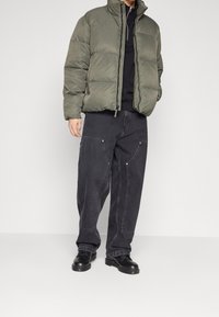 Känslig olivgrön jacka med hög krage, parat med lösa svarta jeans med stora fickor och silverdetaljer, iförd med svarta skor.