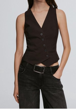 Femme portant un gilet cintré à fines rayures sombres avec des boutons, associé à un jean noir et une ceinture noire, debout avec une main dans la poche.