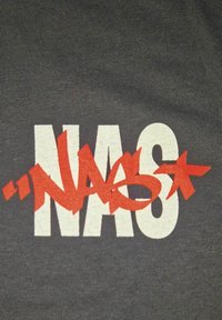 Tmavě šedé tričko s výraznými bílými písmeny "NAS" a červeným textem ve stylu graffiti, který překrývá písmeno "A" a má červenou hvězdičku jako akcent.