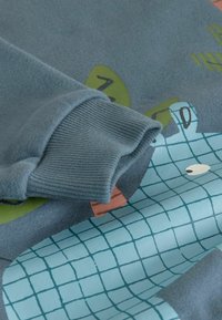 Zila džemperis ar teksturētām piedurknēm, ar rotaļīgu dzīvnieku dizainu un reģipspattem marķējumā maigos, kontrastējošos toņos.