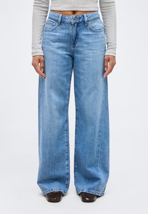 Osoba oblečená v svetlomodrých džínsach s vysokým pásom a širokými nohavicami, v krátkom sivom tričku s dlhým rukávom a hnedých topánkach so štvorcovou špičkou stojaca na bielom pozadí.