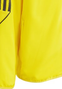 Gelber Sportjacke aus glattem, glänzendem Stoff. Mit schwarzen Streifen und einem strukturierten Taschen-Detail, elastischem Saum am unteren Ende.
