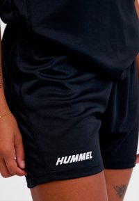 Shorts de sport noirs en tissu léger, dotés d'une ceinture élastique et du logo blanc "HUMMEL" sur le côté. Texture lisse.