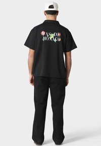 Svart kortermet skjorte med blomstertrykkdesign på baksiden, med teksten "PAS DE MONACO." Kombinert med svarte bukser.