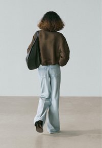 Persoon met krullend haar, gekleed in een bruine trui en lichtblauwe spijkerbroek, loopt weg terwijl hij een grote zwarte schoudertas draagt.