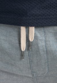 Haut en tissu texturé bleu marine associé à un pantalon en denim bleu clair, doté de cordons crème avec embouts métalliques, mettant en valeur des lignes épurées.