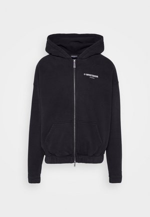 Czarna bluza z kapturem zapinana na zamek błyskawiczny, z przednimi kieszeniami, elastycznym dołem i logo "WRSTBHVR STUDIOS" na lewej piersi, przedstawiona na białym tle.