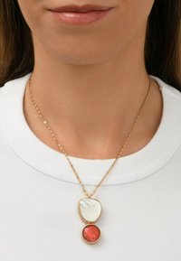 Collier en chaîne dorée avec deux pendentifs : un pendentif supérieur en forme de coquillage blanc lisse et une pierre rouge facettée en dessous. Design simple et élégant.