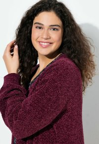 Burgundy-färgad, texturerad kofta med överdimensionerade ärmar, bärs över en blommig topp och har ett mjukt, fluffigt material mot en neutral bakgrund.