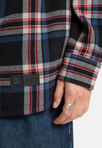Czerwono-niebieska i czarna koszula flanelowa w kratę z plecioną metką na dolnym brzegu. Posiada zapięcie na guziki oraz mankiety z podwinięciem, zaprezentowana na tle dżinsu.