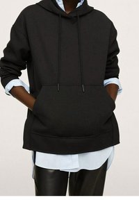 Sudadera negra con capucha, bolsillo frontal, cordones y costuras de hombro caídas, sobre una camisa azul claro y pantalones negros.